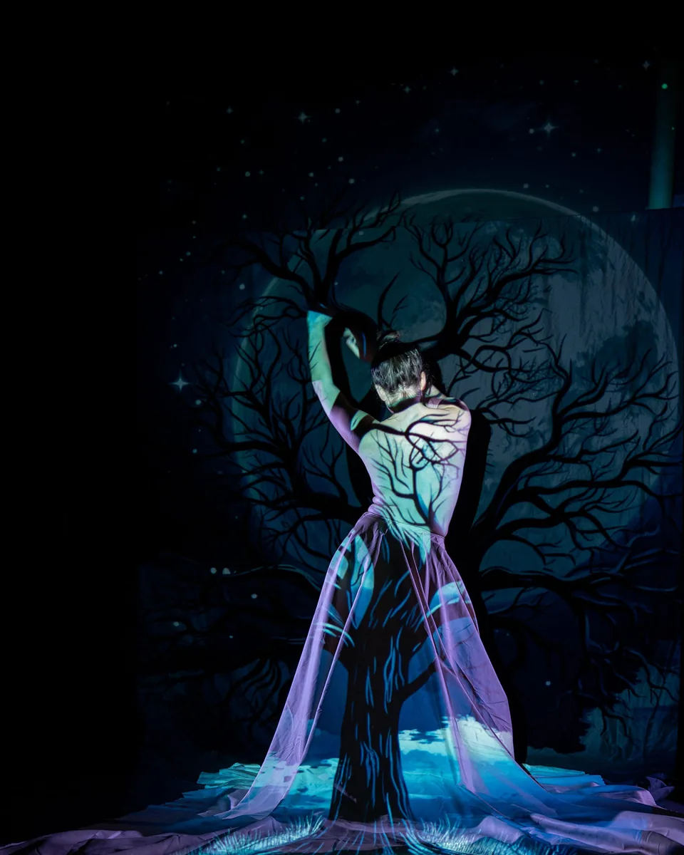 A figure stands with arms raised, illuminated by cool blue and purple light with a projection of a tree and branches covering their bare back and flowing fabric skirt, set against a dark starry backdrop.