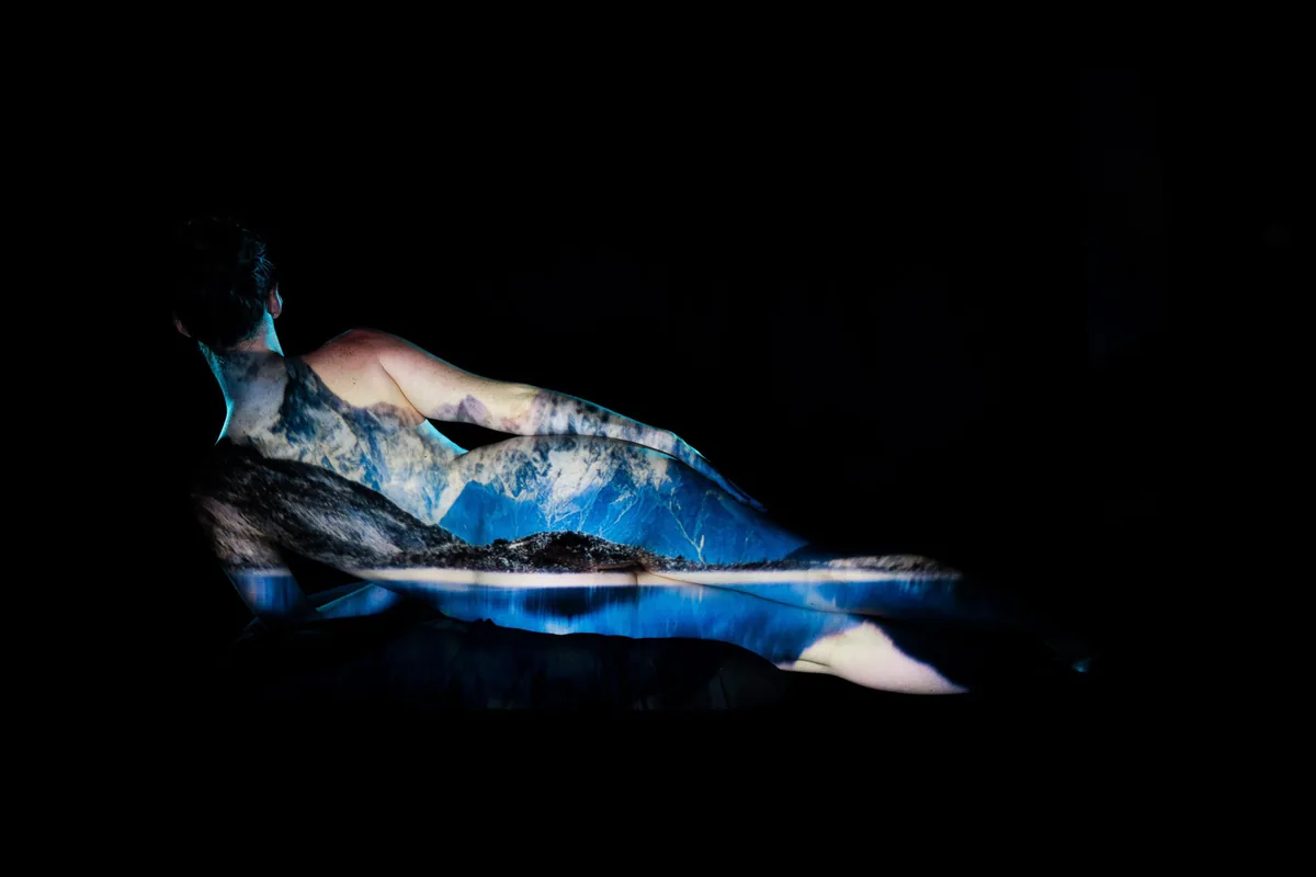 A figure reclines against a dark background with a cool-toned mountain landscape projected onto their skin, the relaxed pose emphasizing gentle curves and quiet stillness.