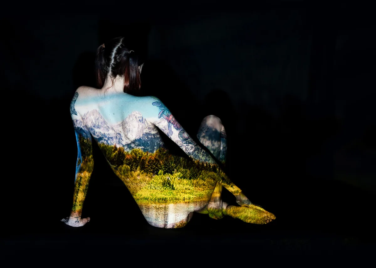 A seated figure with back turned, draped in a projection of a natural landscape featuring mountains, trees, and water, the dark background emphasizing the projected light and colors.