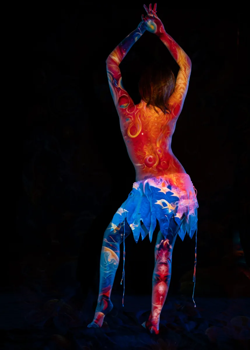 A person stands barefoot with arms raised above their head, light projections in red, blue, and yellow swirling patterns cover their back, arms, and legs, against a dark background.