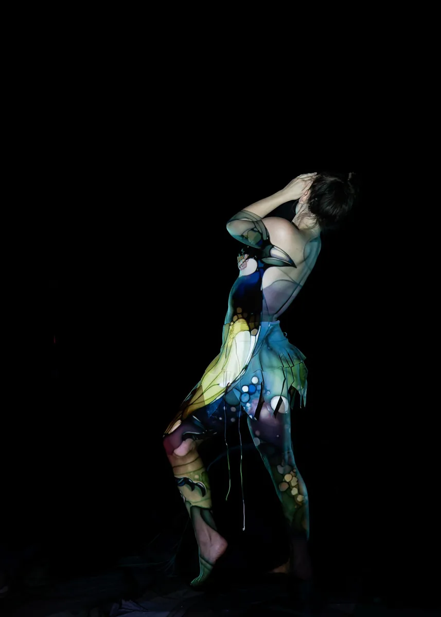 A person stands with their back slightly arched and hands covering their face, with a stained glass-style projection in blue, green, and yellow hues cast over their body and a sheer fabric skirt.