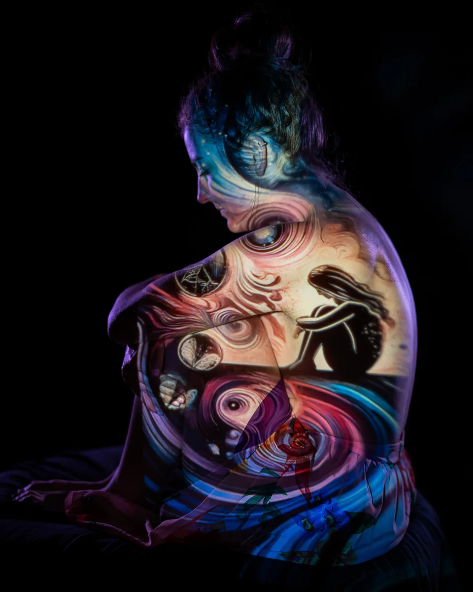 A woman sits curled with her knees drawn up, illuminated by a swirl of warm, colorful projected artwork depicting a seated figure and abstract shapes over her skin and fabric, set against a dark background.