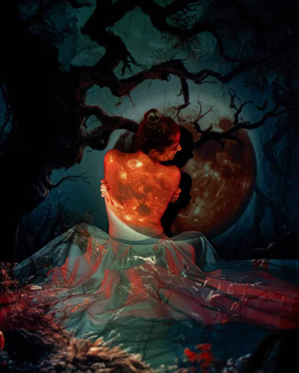 A woman seated with her back to the camera, arms crossed gently, illuminated by warm orange and red light of a moon projection, wearing a flowing translucent fabric with a dark tree silhouette in the background.