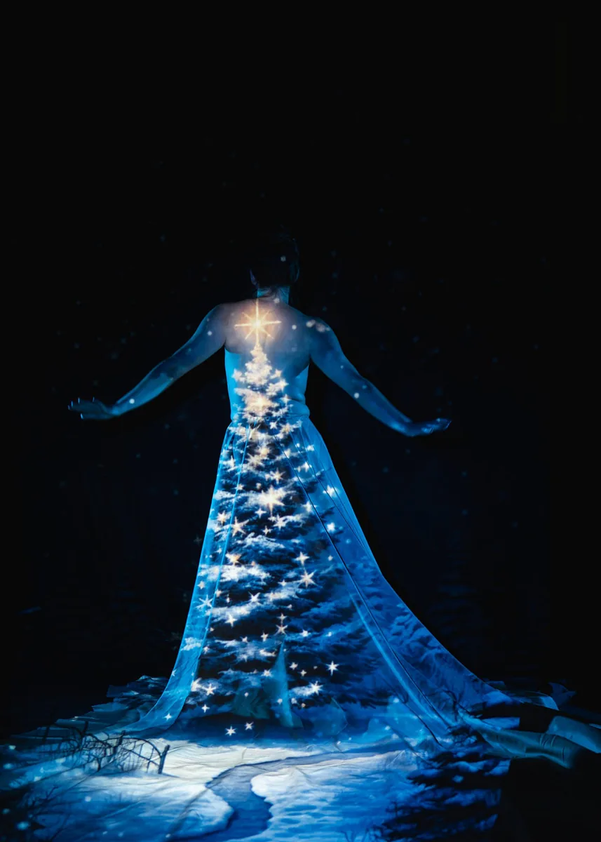 A figure stands with arms extended as a blue-toned winter scene, featuring a decorated tree with a glowing star on top, is projected onto a flowing fabric dress, set against a dark background.