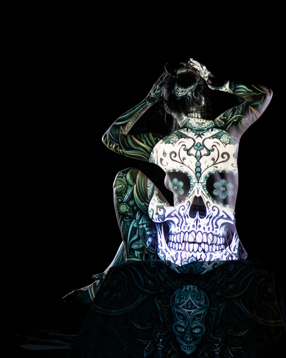 A person sits with one knee raised and hands resting on the head, while an intricate skull and floral pattern is projected across their back and limbs, set against a dark background.