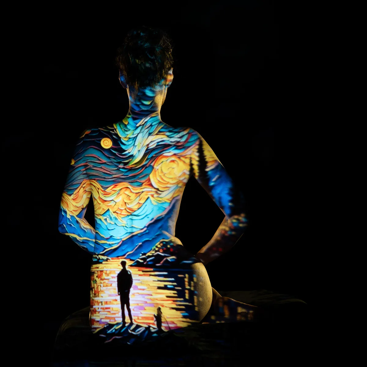 A person sits with their back to the camera in darkness, a vibrant projection of a colorful evening sky and a silhouette by water covering their skin and casting warm and cool tones.