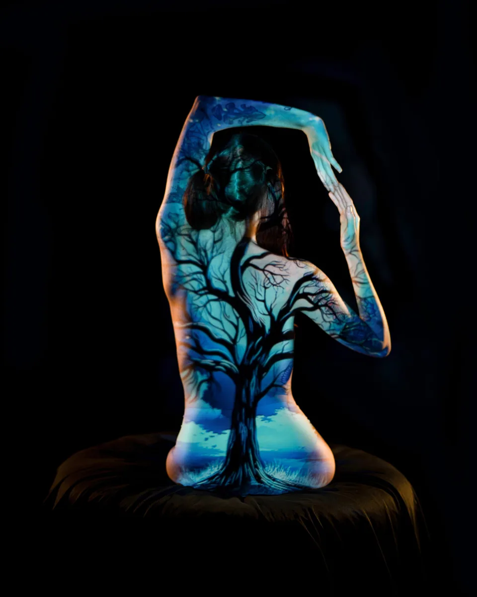 A person is seated with their back to the camera, arms arched above the head while a vivid blue and black tree is projected onto their bare skin, set against a dark background.
