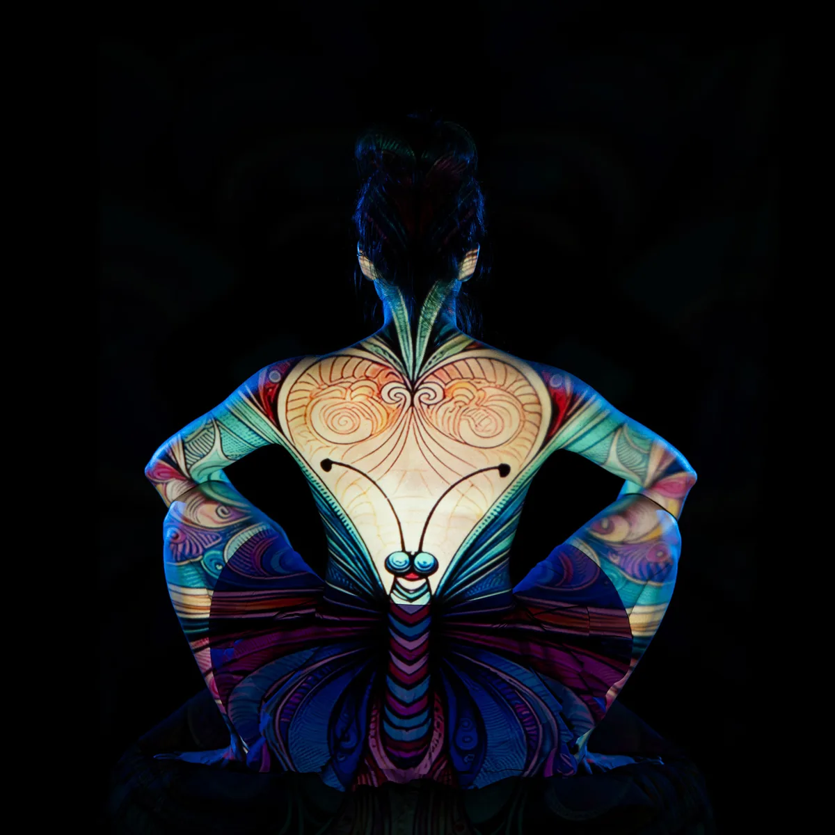 A person sits cross-legged with their back to the camera, illuminated by colorful butterfly artwork projected across their bare skin, creating a layered and contemplative mood.