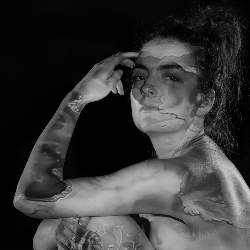 Nude person with hair tied up sitting with arm raised, textured shadows with abstract patterns cast on face and body against a black background