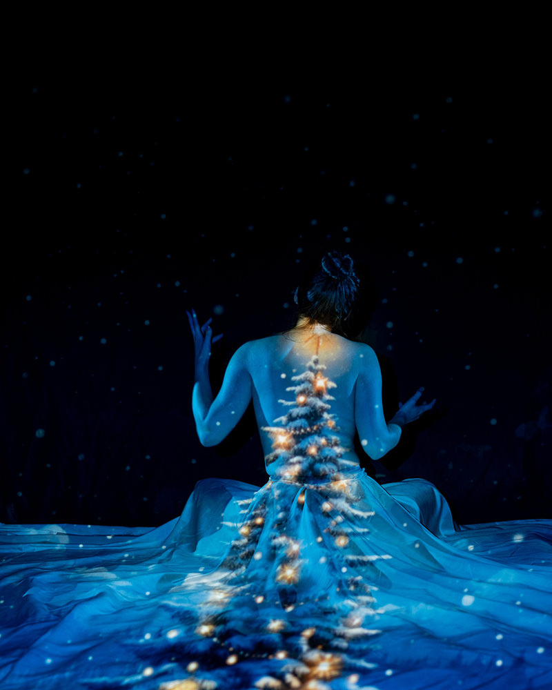 Seated person facing away with a Christmas tree with glowing lights projected onto their back and extending onto the fabric around them, surrounded by small light specks resembling snowflakes against a dark background