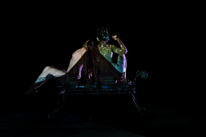 Two figures seated back-to-back on a bench with overlapping textures and patterns projected onto their bodies against a black background