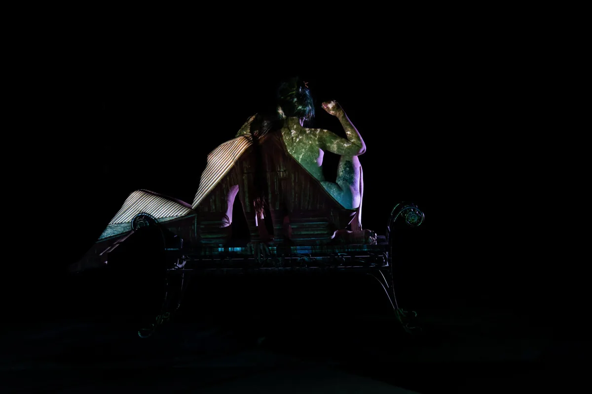 Two figures seated back-to-back on a bench with overlapping textures and patterns projected onto their bodies against a black background