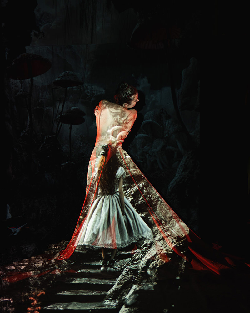 Woman with hair in a bun turned to the side, with projection of a girl in a dress walking on stone steps in a dark, fantasy-like forest with large mushrooms and textured rocks surrounding.