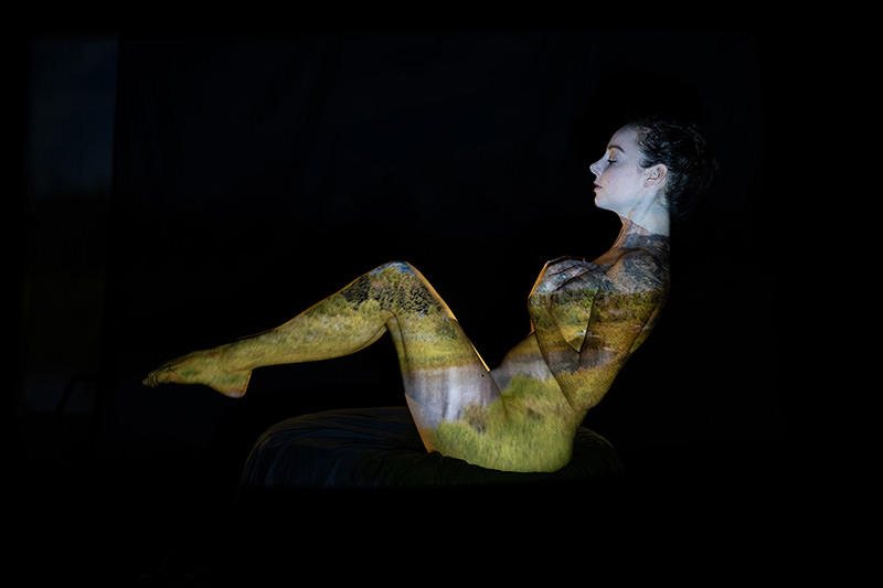 Side view of a nude woman sitting with knees bent and arms crossed over her chest, with a landscape of green trees and grasses projected onto her body against a black background