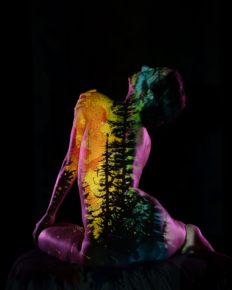 A person with their back to the camera, sitting and leaning back, with a colorful projection of trees and a vibrant yellow-orange sky cast over their body against a dark background.