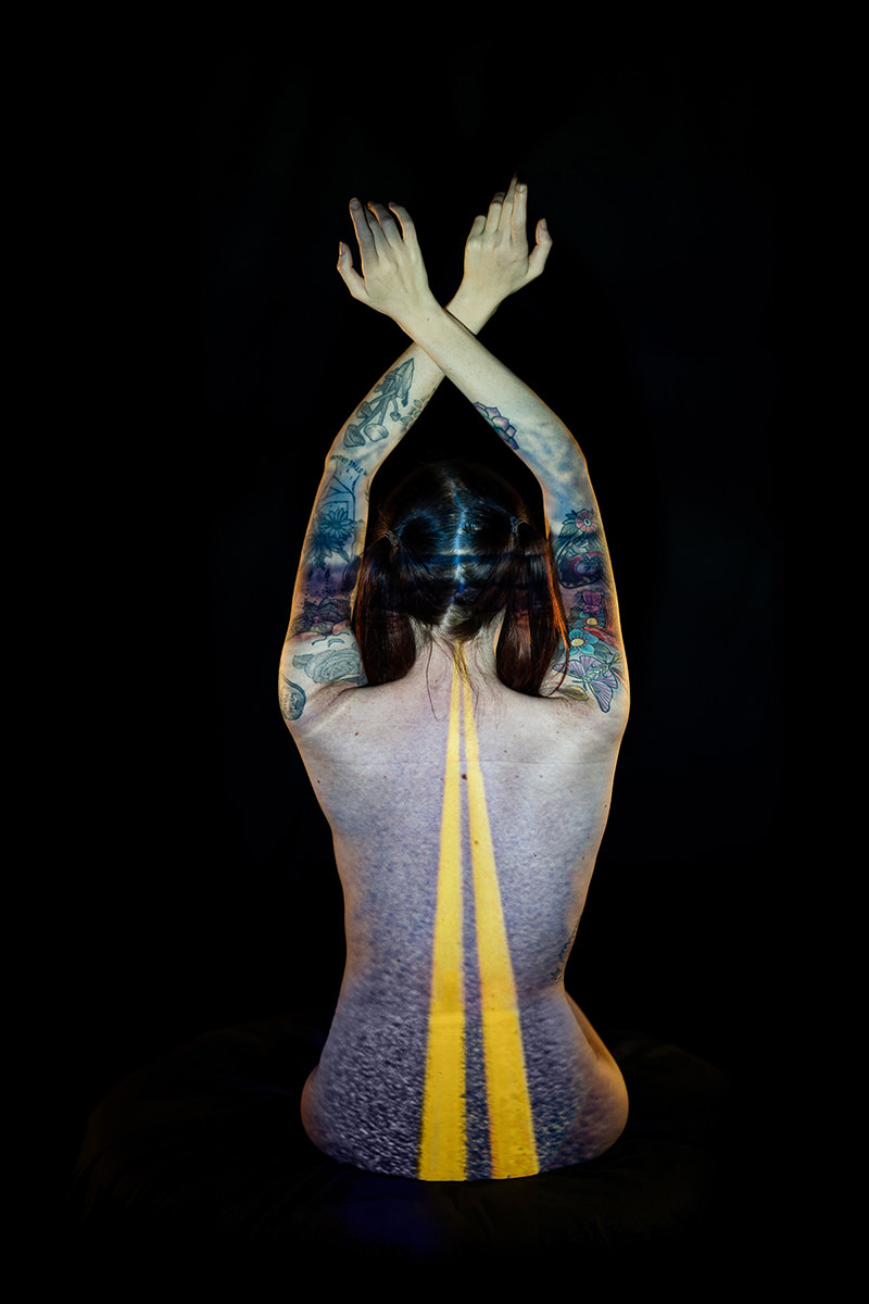 Person seen from the back with arms crossed overhead, covered in detailed colorful tattoos, with a projection of a road featuring double yellow lines running vertically down their back against a black background