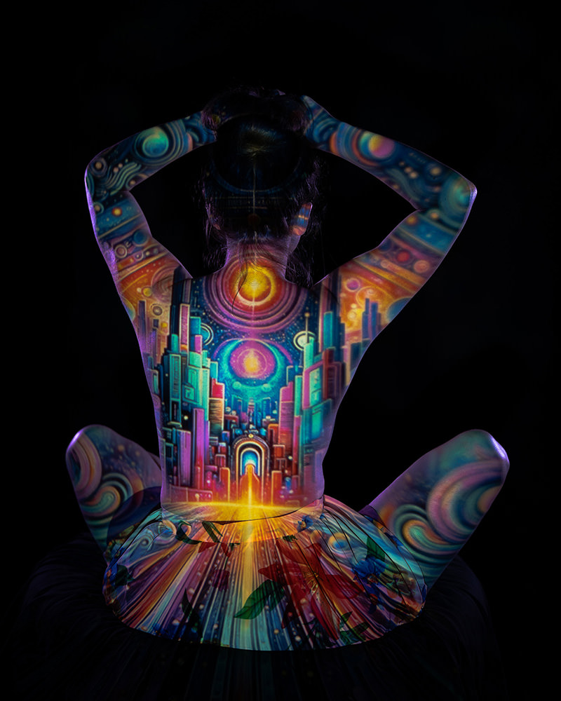 Back view of a seated dancer with arms raised, displaying a colorful projection of a futuristic cityscape with bright lights and abstract patterns on her skin and skirt against a black background