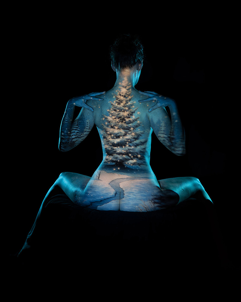 Rear view of a person seated with knees bent, with a projection of a snowy winter scene featuring a decorated Christmas tree and a snowy path cast on their back and arms against a dark background