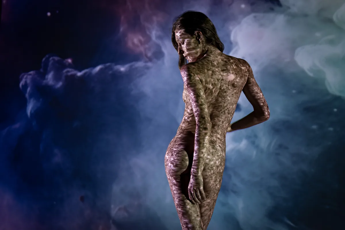 A person stands with their back slightly turned, their bare skin covered by textured light patterns in a dark space against a backdrop of diffuse purple and blue shapes.