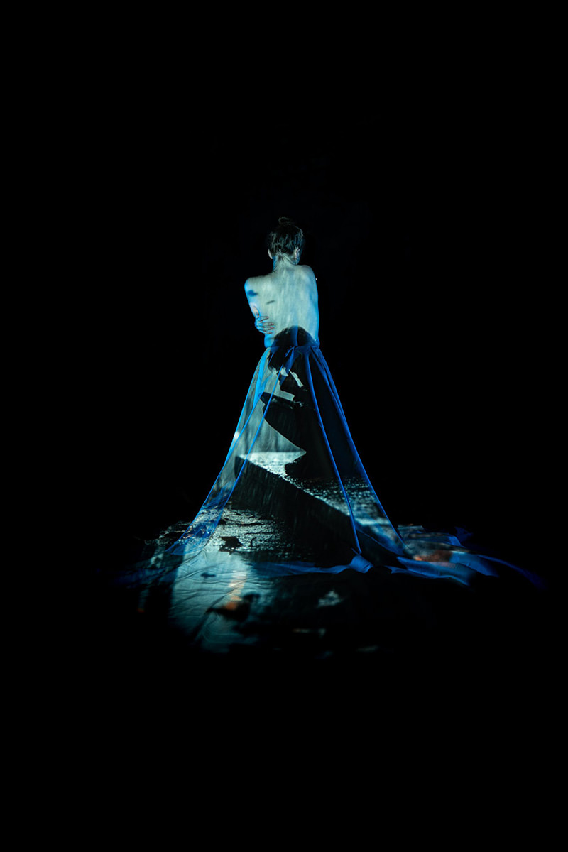 A person with bare back facing away, wrapped in a flowing translucent fabric with a projection of a coastal scene featuring a stone wall and water, on a black background.