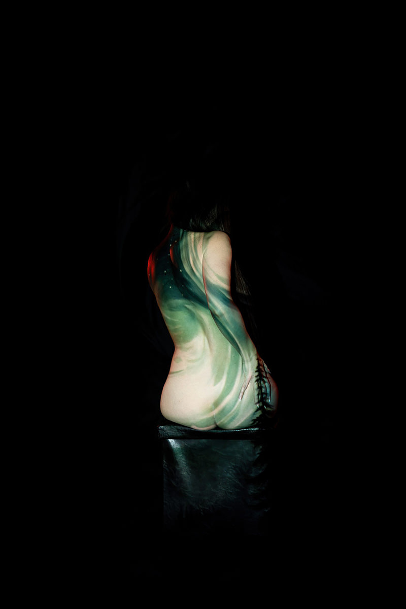 Nude person sitting on a black stool with back facing the camera, body painted with green and blue flowing abstract patterns, set against a dark background