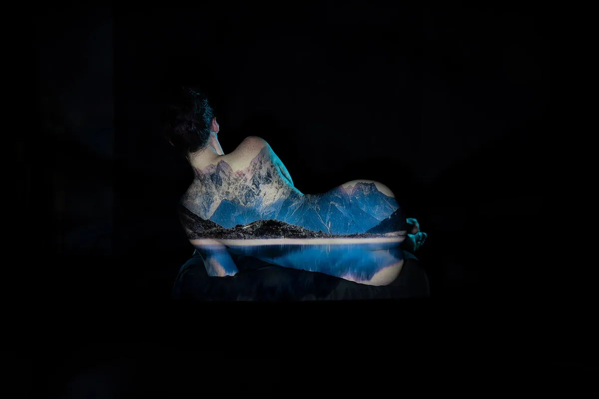 Nude person lying on side with a detailed mountain and lake landscape projected onto their back and legs against a black background