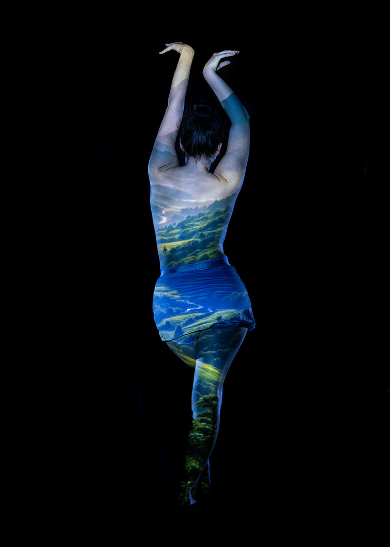 Person standing on one leg with arms raised, back facing viewer, a vivid landscape scene of hills and trees projected over their body against a black background