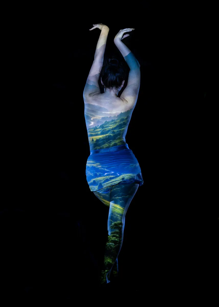 Person standing on one leg with arms raised, back facing viewer, a vivid landscape scene of hills and trees projected over their body against a black background