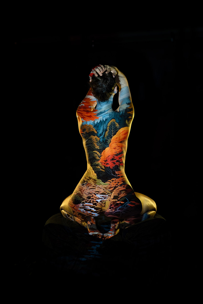 Person seen from the back seated with arms raised and hands clasped behind the head, with a vibrant autumn landscape including trees and a winding river projected onto their body, set against a black background