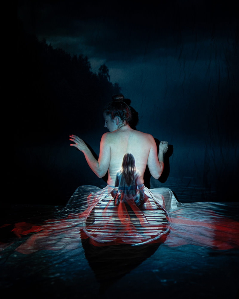 Back view of a woman sitting topless with a projection of a path and a smaller figure of a girl sitting on her back, set against a dark, moody background
