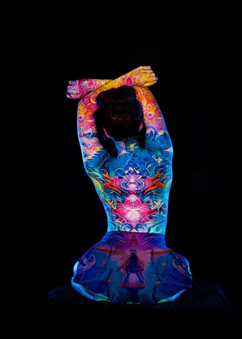 Person seen from the back with colorful, intricate tattoos covering the shoulders, upper back, and arms, against a black background. The individual has their arms raised and crossed behind their head, with the tattoo art featuring vivid blues, pinks, and oranges in a symmetrical design.