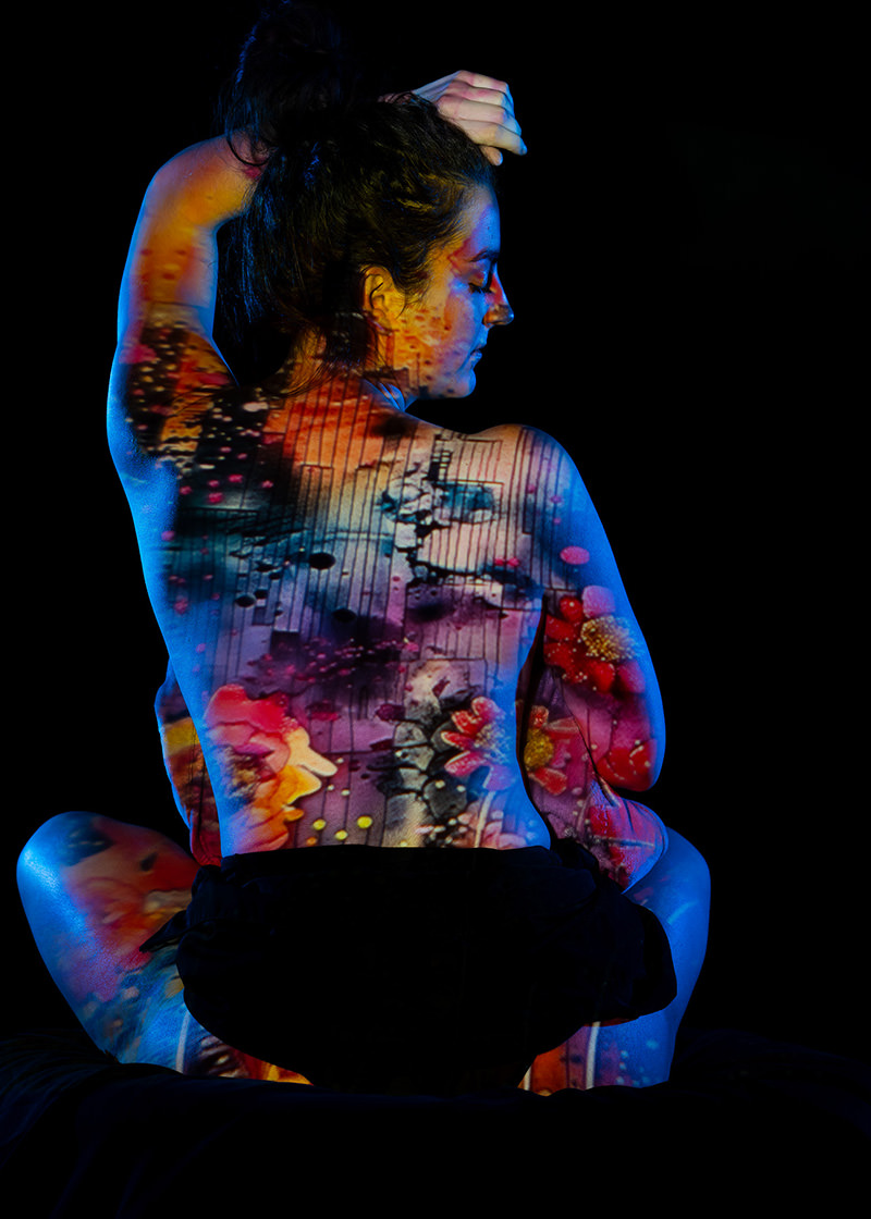 Person sitting cross-legged with their back facing the viewer, head turned sideways and one arm raised, covered in a vibrant, colorful projection of abstract floral and geometric patterns against a black background