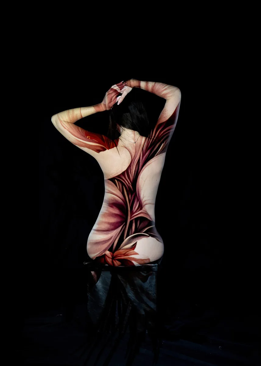 Back view of a seated person with arms raised, covered in intricate floral body paint blending shades of red, maroon, and beige, against a black background