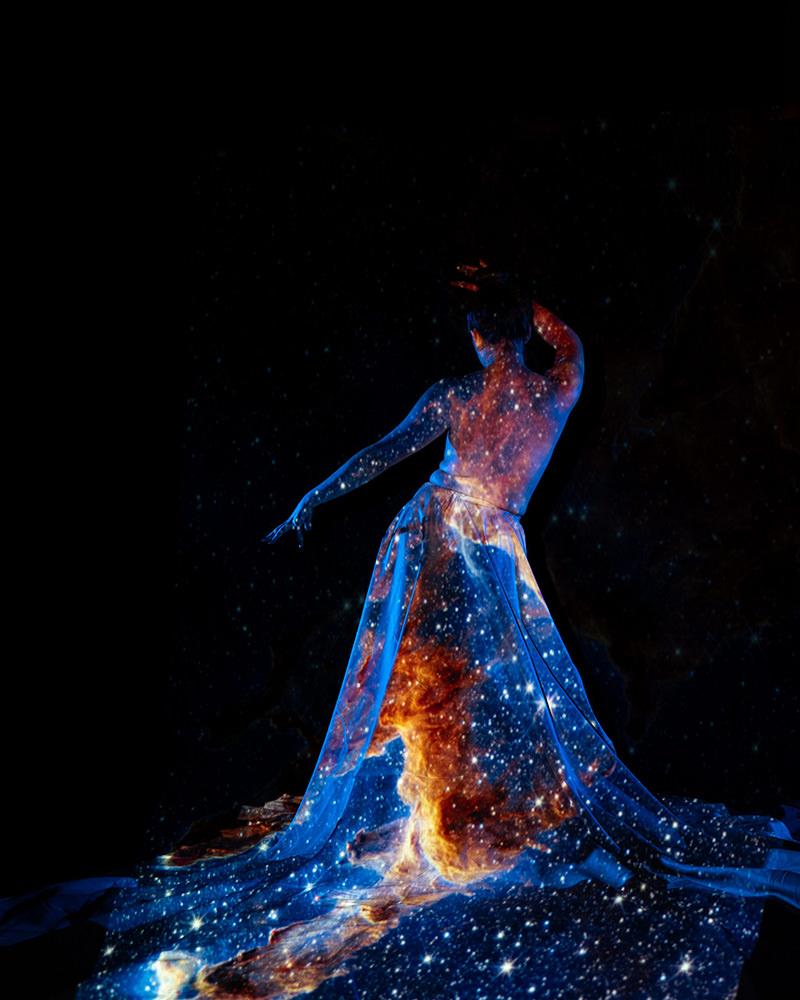 A woman with her back to the camera, draped in a flowing fabric covered with a projection of a vibrant, star-filled galaxy against a dark background.