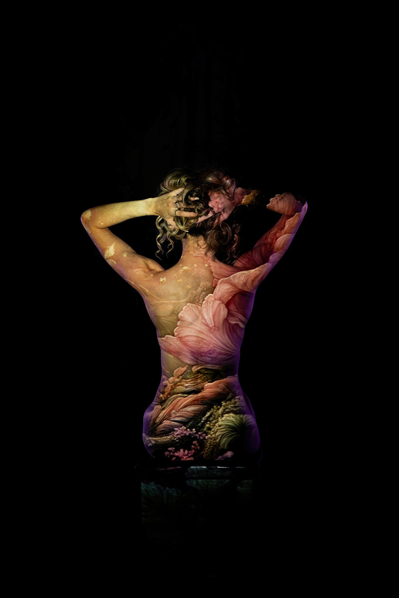 A woman with curled hair seen from behind, holding her hair up with both hands, her bare back illuminated with a projection of large pink flowers and green plants against a black background.