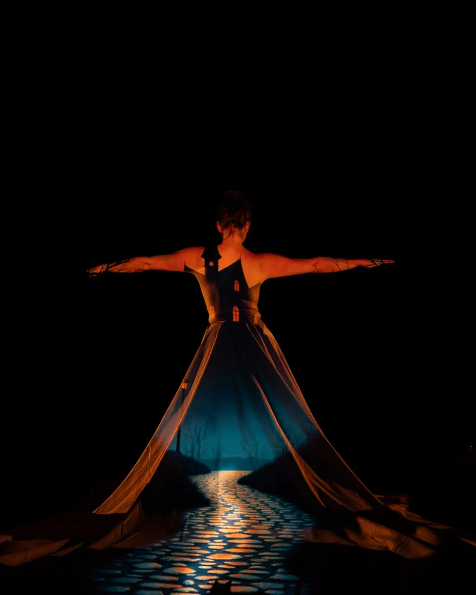 Person standing with arms extended wearing a long flowing dress with a projection of a cobblestone path leading to a haunted house and barren trees on the back of the dress and upper body against a dark background