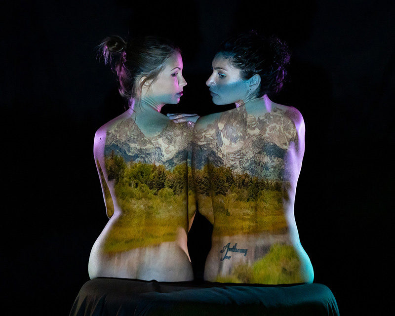Two women sitting back-to-back with a mountain and forest landscape projected onto their bare backs, set against a dark background