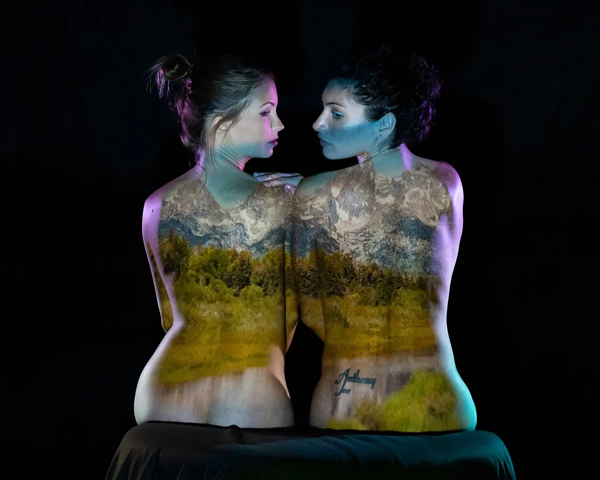 Two women sitting back-to-back with a mountain and forest landscape projected onto their bare backs, set against a dark background
