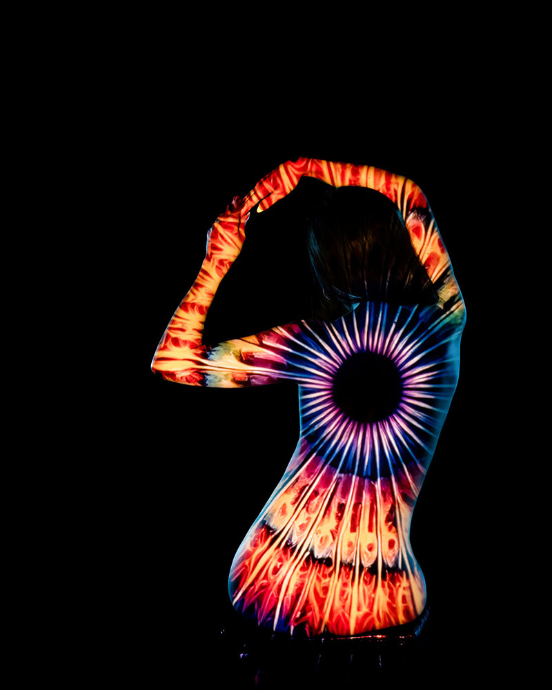 Person with arms raised above head, body illuminated by colorful psychedelic tie-dye pattern, set against a black background