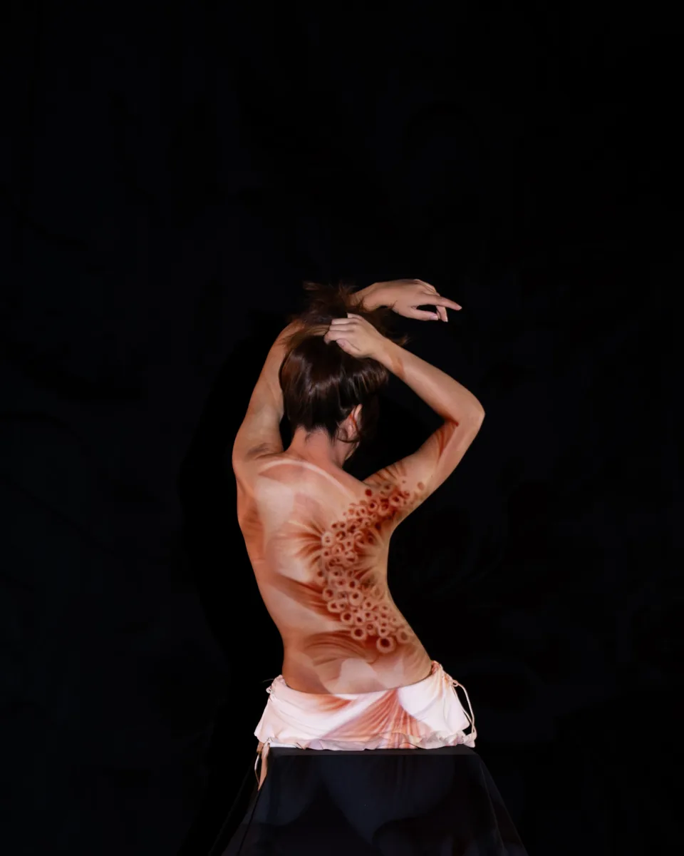 A person seen from behind with light projection of organic, circular patterns in warm tones on their back, set against a dark background, with arms raised to hold their hair.