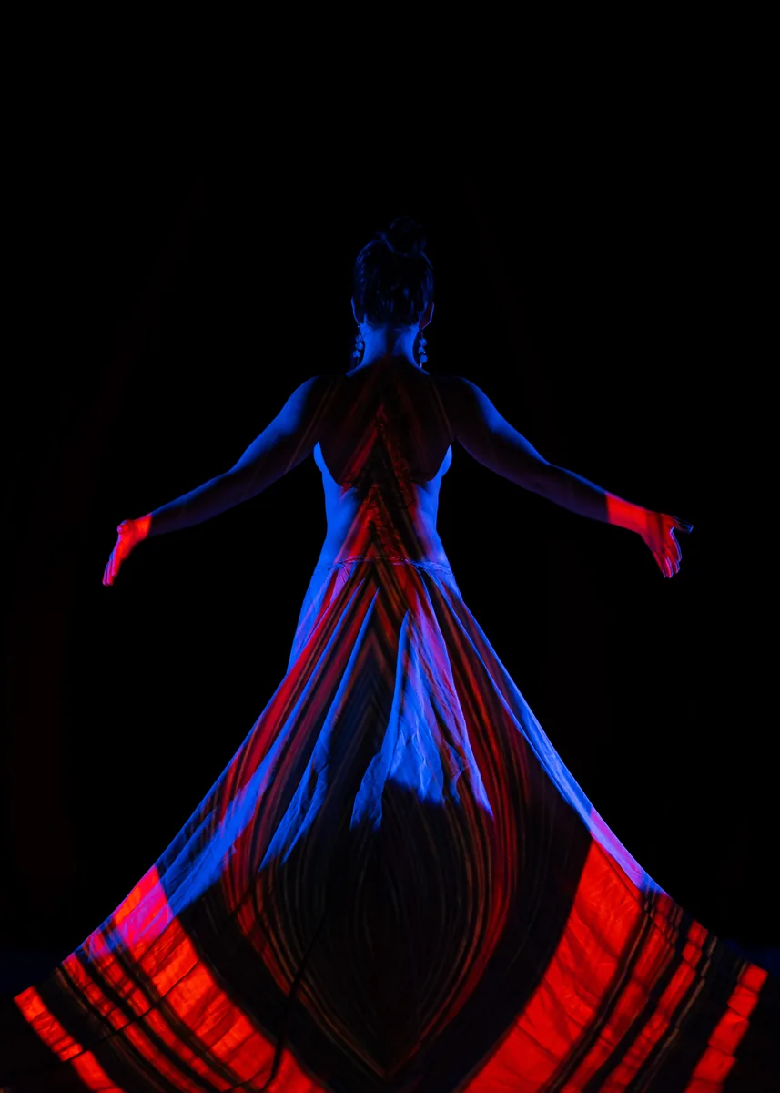 A person stands with their back to the camera, arms extended, light projecting red and blue geometric patterns across their back and flowing skirt against a dark background.