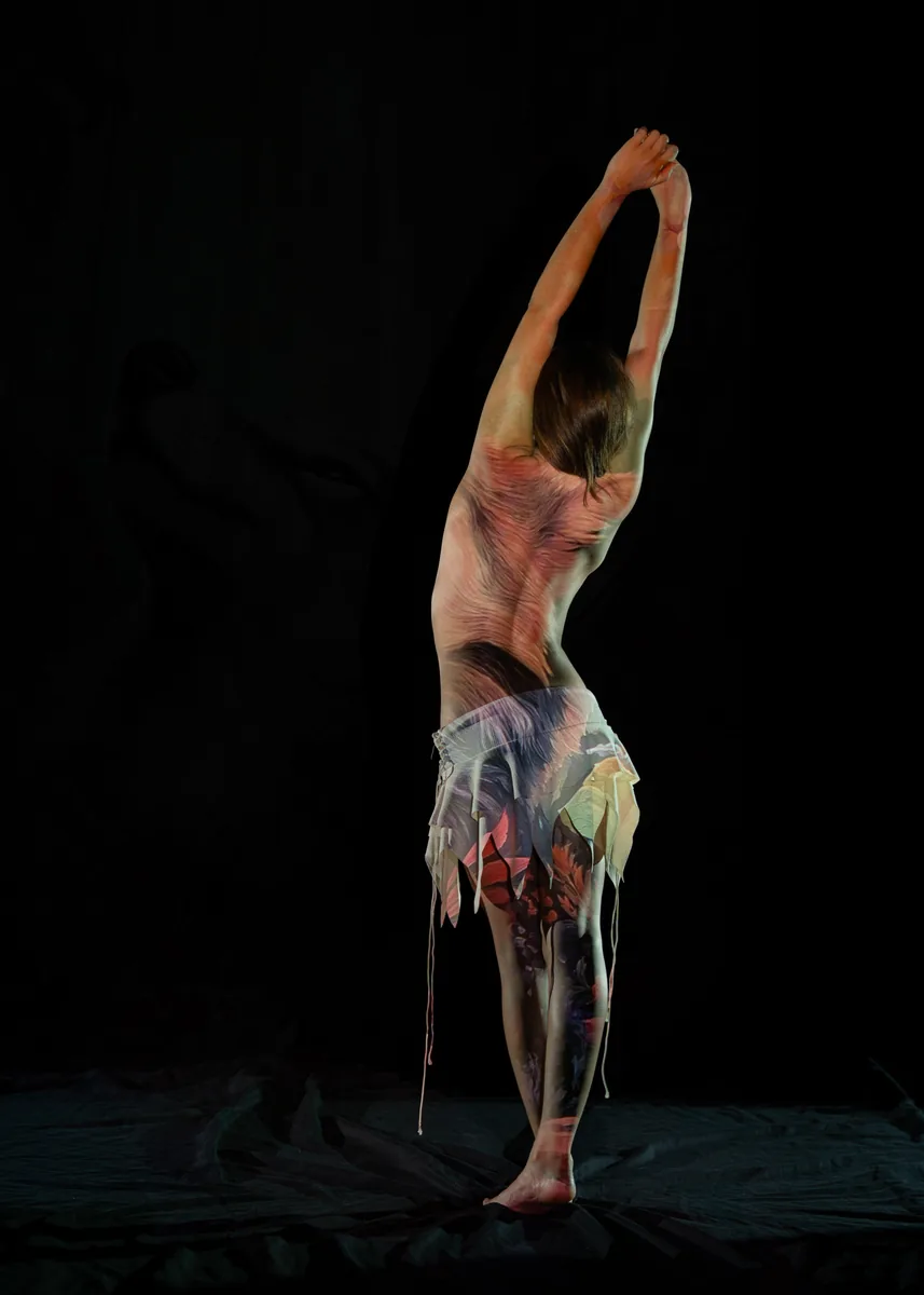 A person stands on one foot with arms raised and hands clasped above the head, light projections of colors and designs cover their back and the sheer fabric worn around their waist.