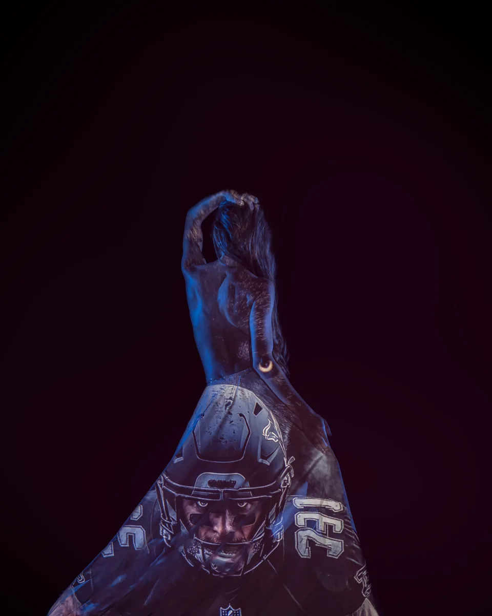 A person with long hair is posed with one arm raised, while a dark image of a football player with a helmet is projected onto their back and lower body against a dark background.