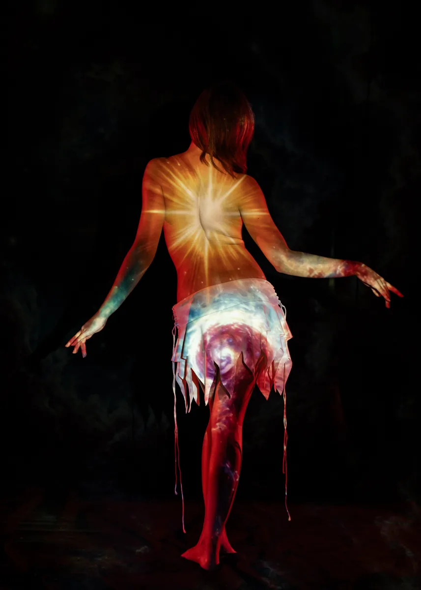 A person is standing with their back to the camera, arms extended slightly, with light projecting a bright star and swirling galaxy pattern on their skin and white skirt against a dark background.