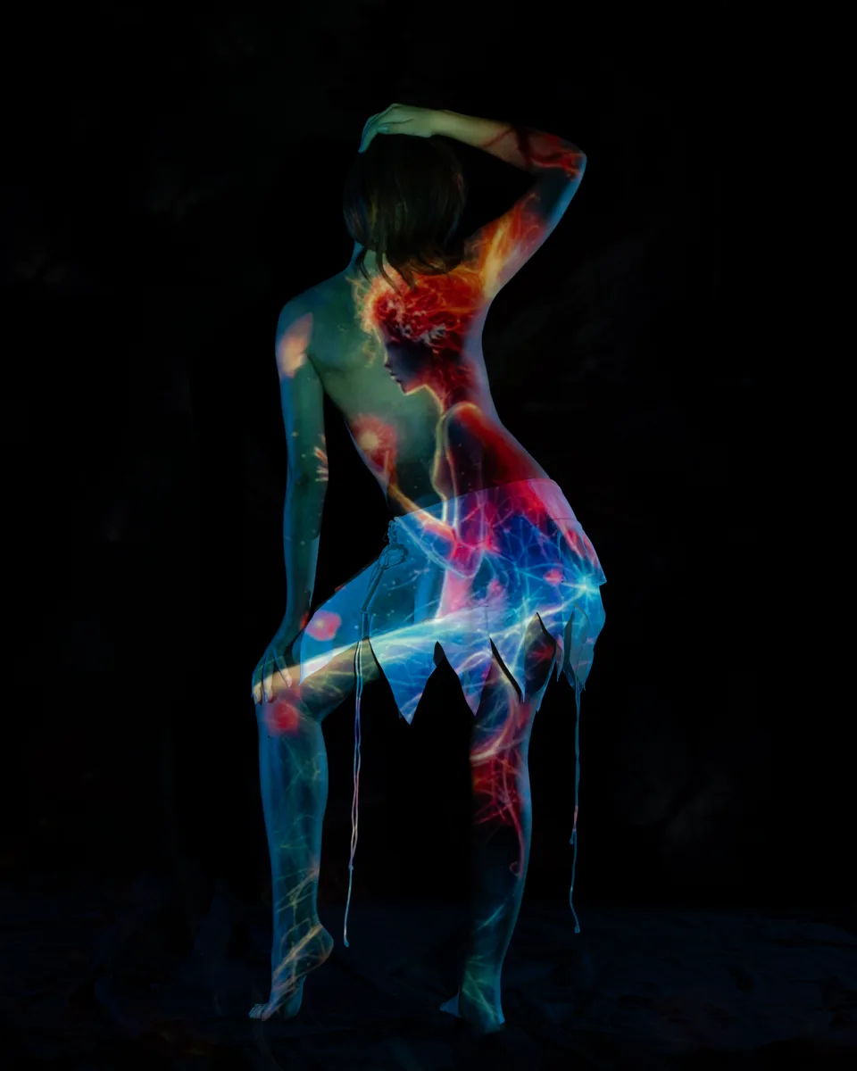Back view of a person standing on one leg with one arm resting on their head and the other on their knee, illuminated by a colorful projection of layered abstract and figurative light patterns against a dark background.