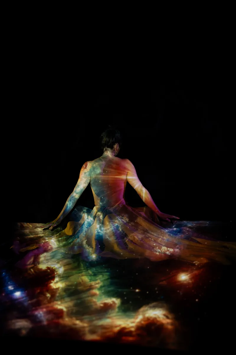 A person sits with their back to the camera, light projecting a vivid, multicolored nebula pattern onto their bare skin and flowing fabric, set against a dark background.