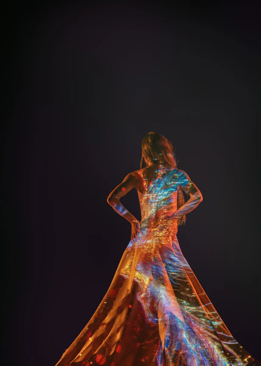A person stands with their back to the camera in a dark studio, light and colorful abstract patterns projected across their bare back and long flowing skirt.