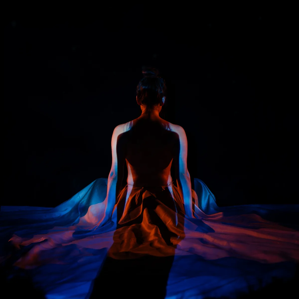 A person sits with their back to the camera, draped in translucent fabric, illuminated by light in warm orange and cool blue tones against a dark background.