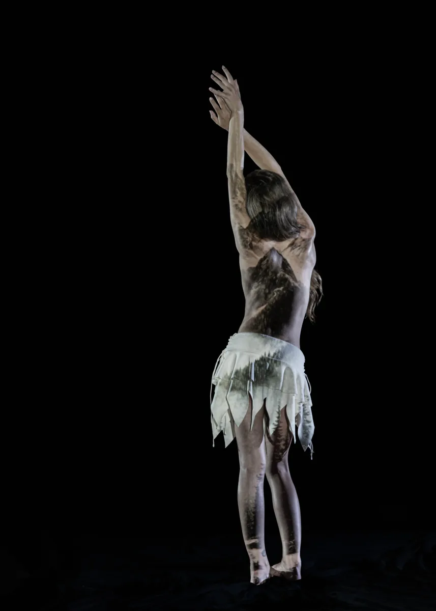 A person stands with their back to the camera, arms extended upward, with a projection of a mountain and forest trees displayed across their back and legs, wearing a white skirt against a dark background.