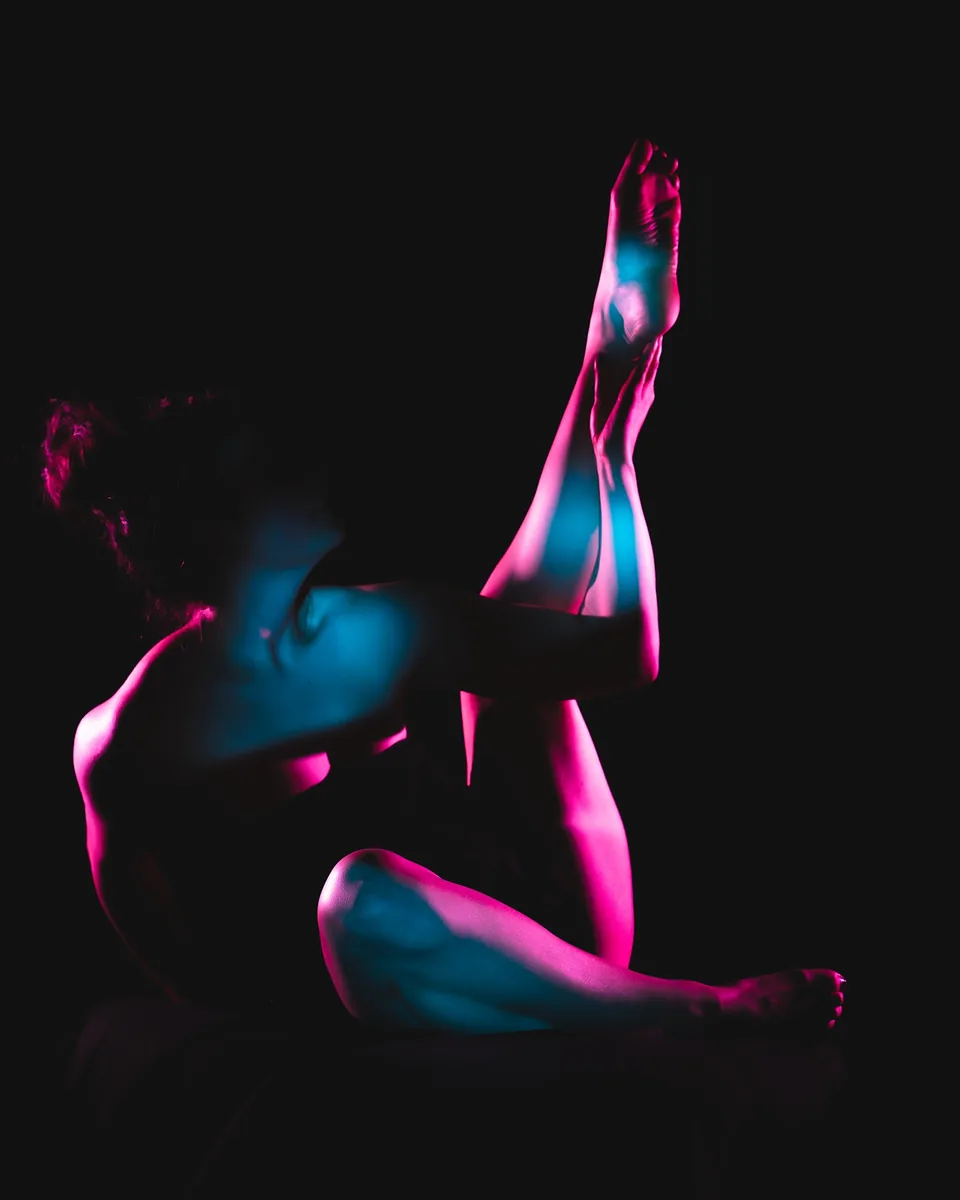 A person sits against a black background with blue and pink light cast across their body, holding one leg extended upwards while partially draped in dark fabric.